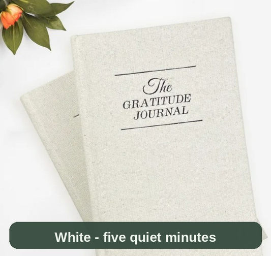 The Gratitude Journal in a white linen finish, resting beside soft greenery - five quiet minutes to end the day.